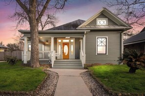 A two-story house with a front porch, a central entrance door, and a small front yard with a tree and a palm plant. - Southtown Charm: Pool, King Beds, Arcade & Games (San Antonio)