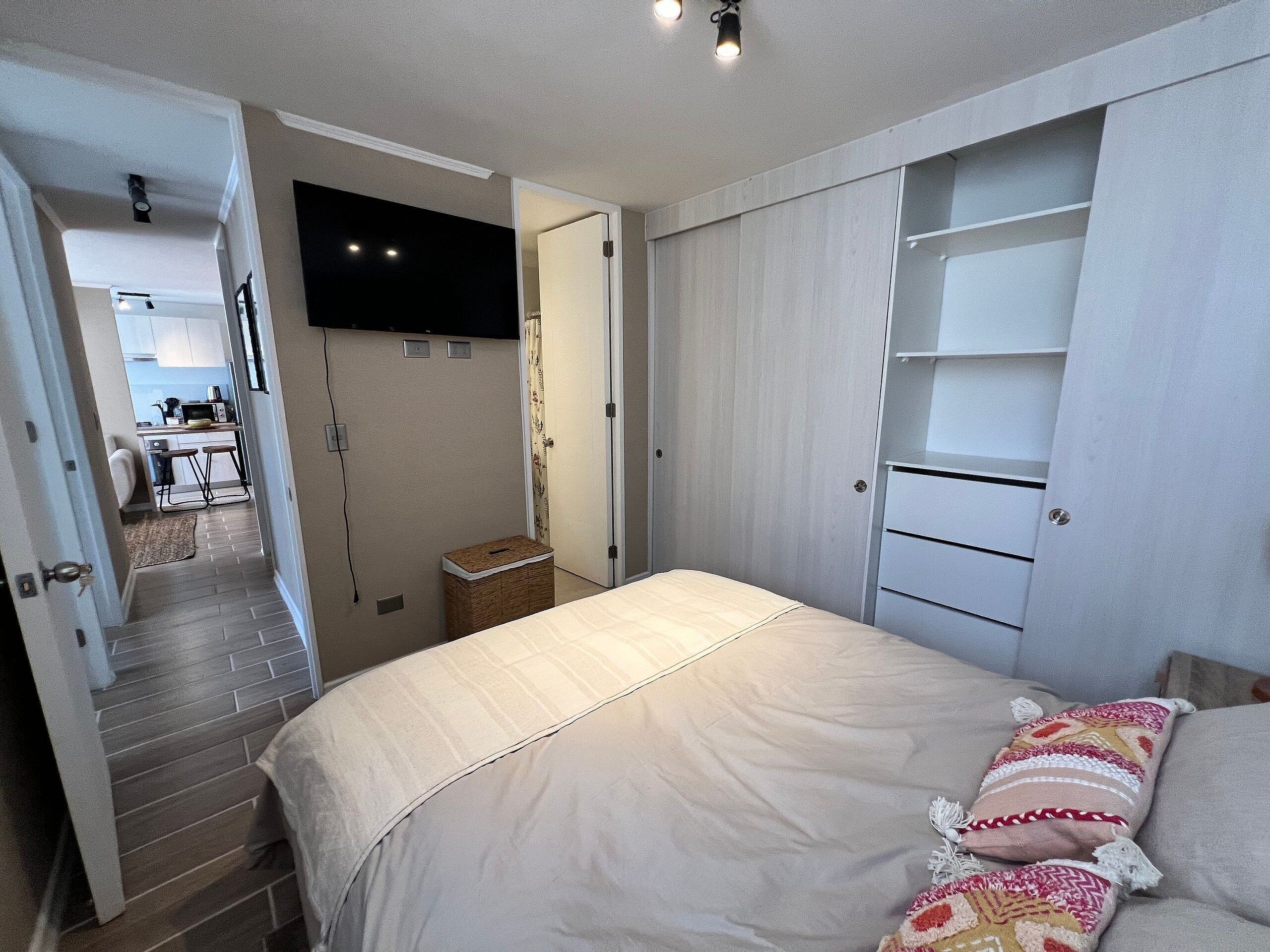 A bedroom with a bed, a TV mounted on the wall, a wardrobe, and a small table with a lamp.