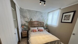 A bedroom with a bed, a nightstand, a lamp, a wall with a tropical mural, a window, and a door. - Oceana Suites Bosque Pucalan (Puerto Montt)