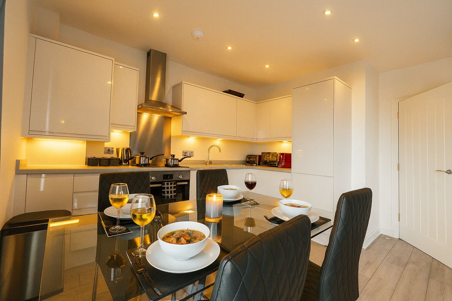 Een moderne keuken met witte kasten, een roestvrijstalen afzuigkap, en een eethoek met een glazen tafel voor twee personen.