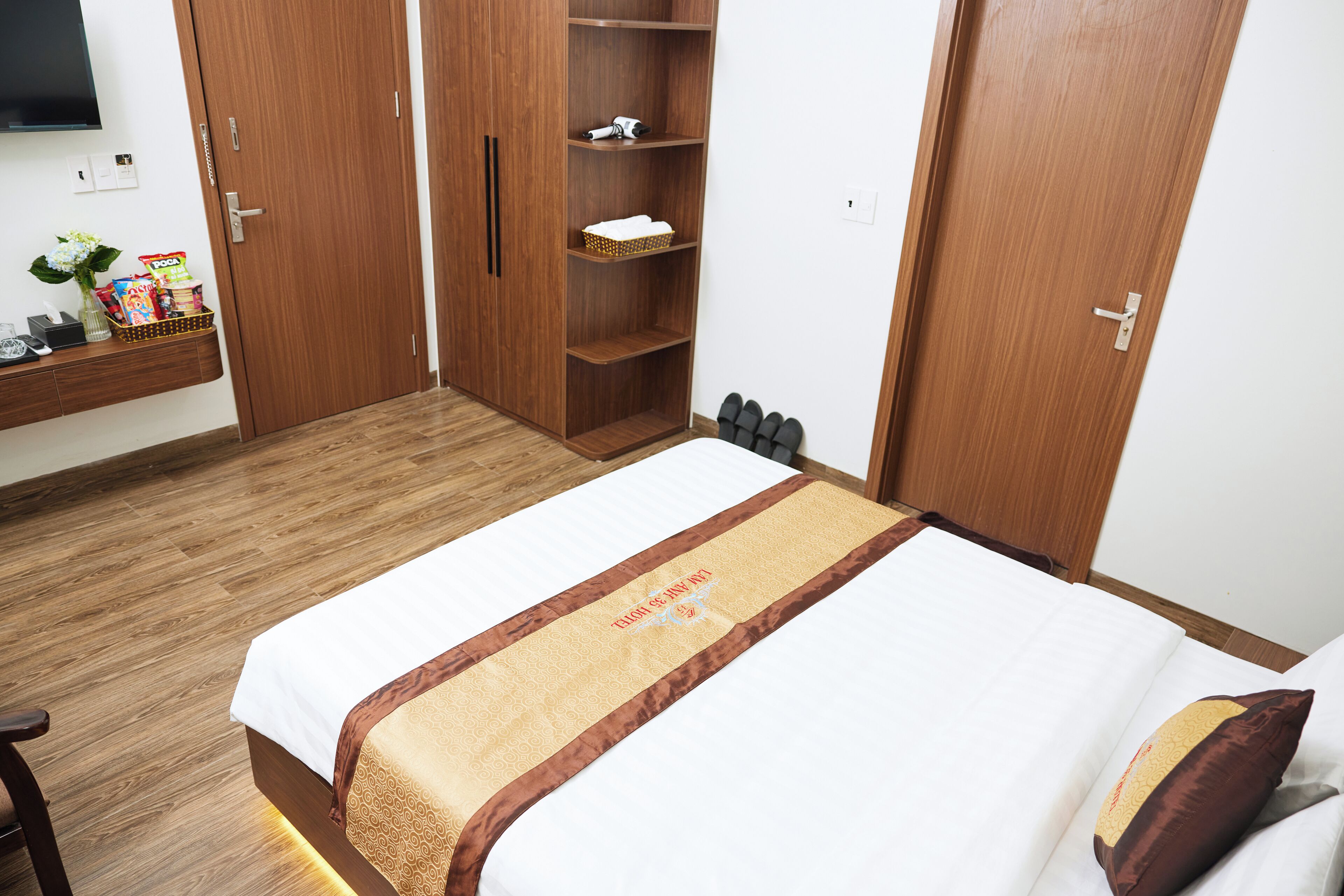 A hotel room with a bed, a wooden wardrobe, a shelf with toiletries, and a small table with a vase and snacks.
