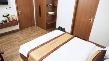 A hotel room with a bed, a wooden wardrobe, a shelf with toiletries, and a small table with a vase and snacks.