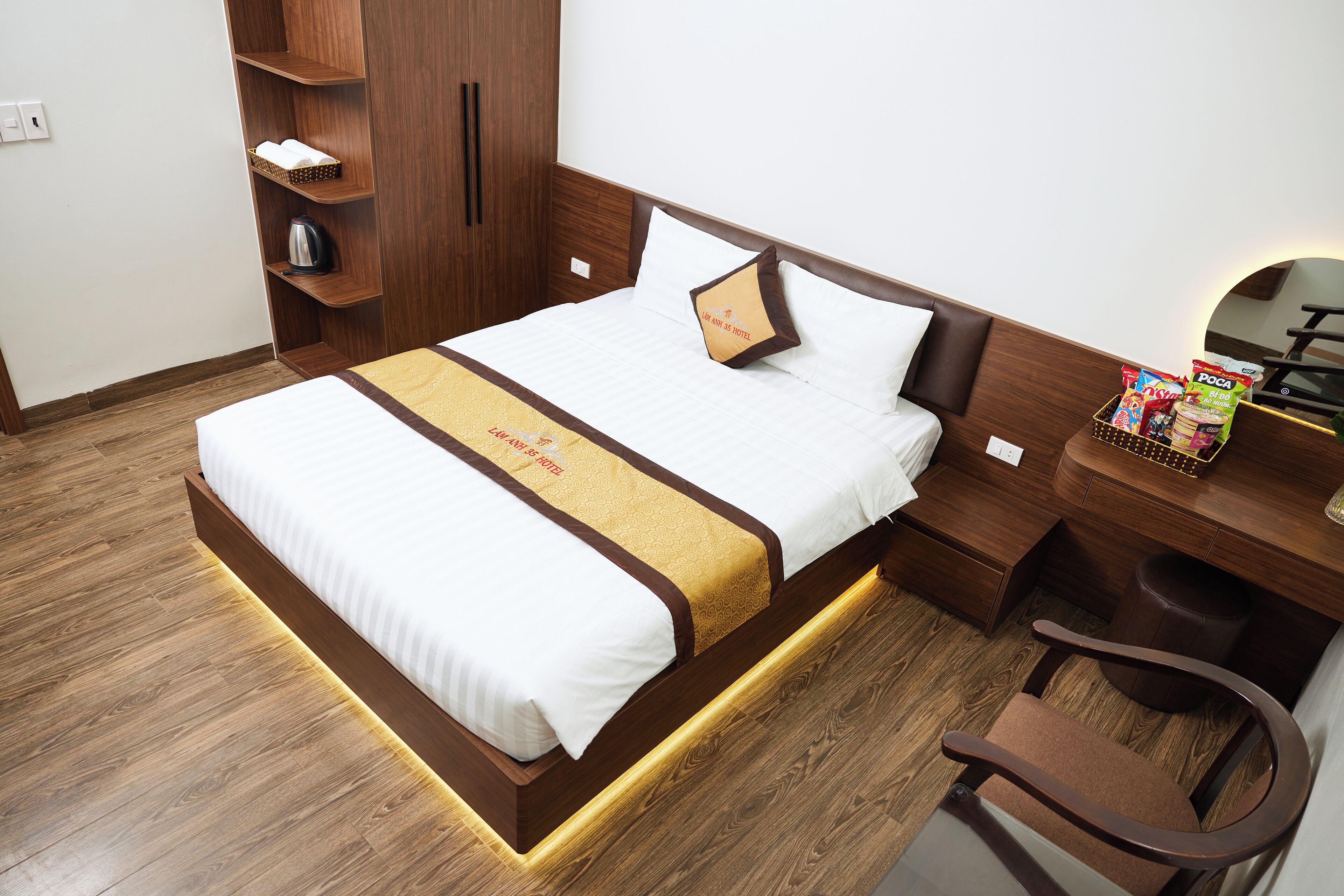 A neatly made bed with a white and gold bedspread, a wooden nightstand with a chair, and a shelf with a kettle and a basket.