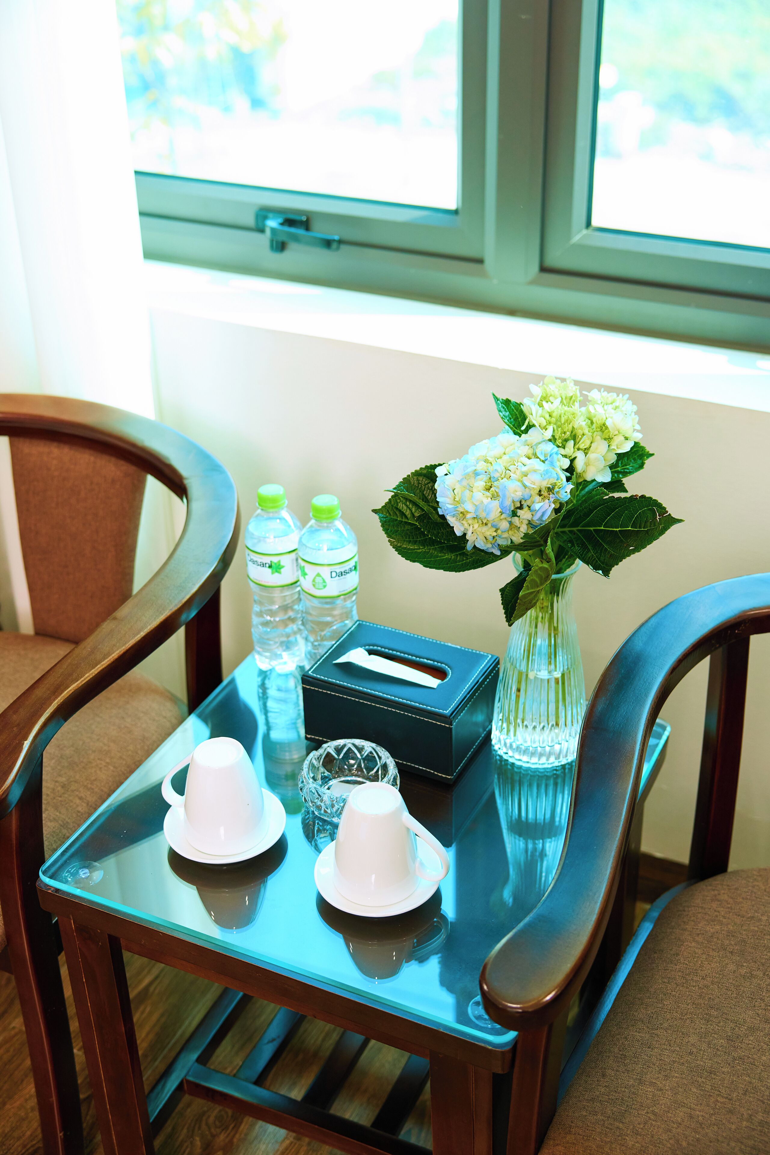 Une table en verre avec deux chaises, un vase avec des fleurs, deux bouteilles d’eau, une boîte de mouchoirs et deux tasses blanches.