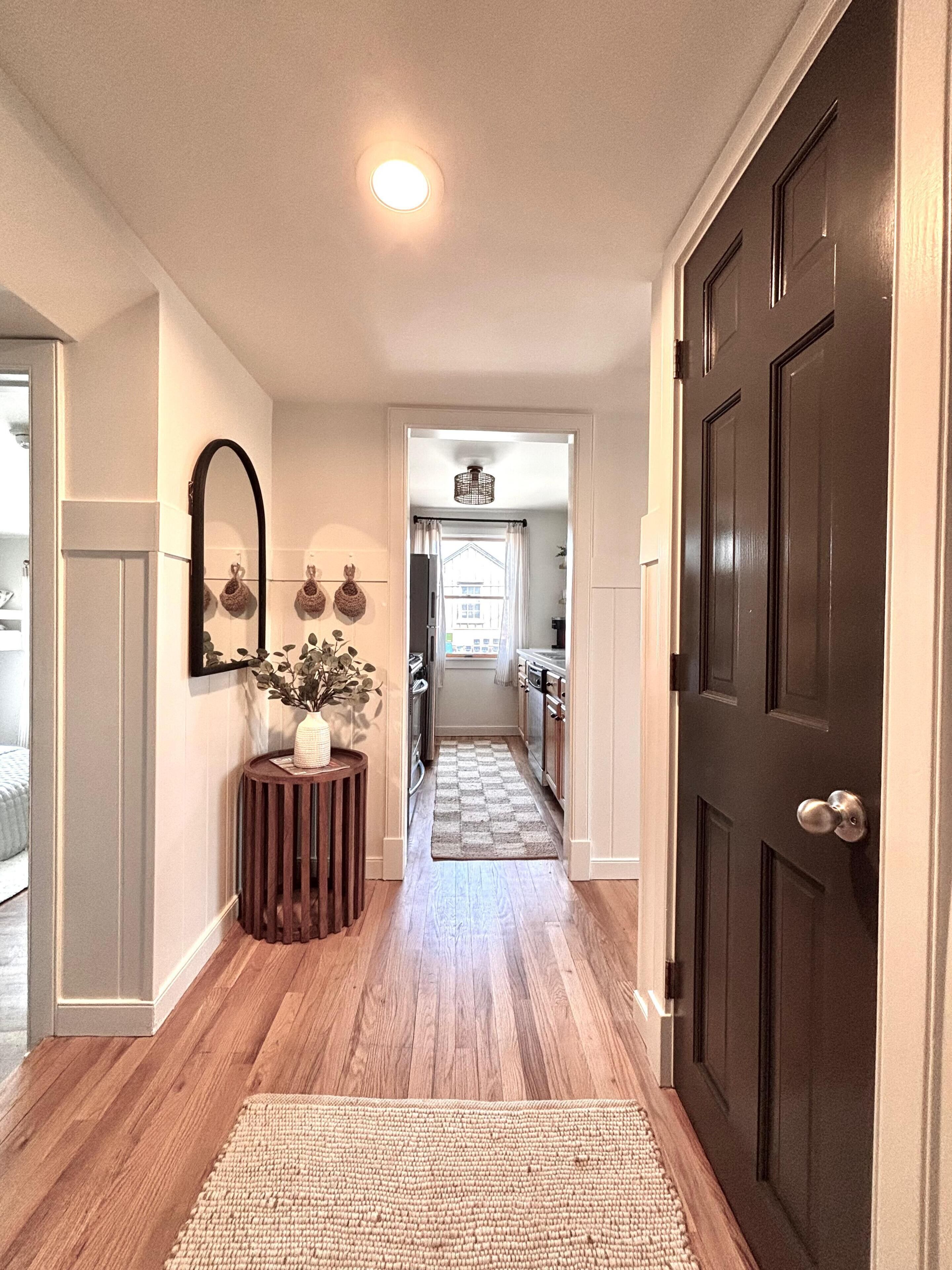 A hallway with wooden flooring, a dark door, a mirror, and a small table with a vase.