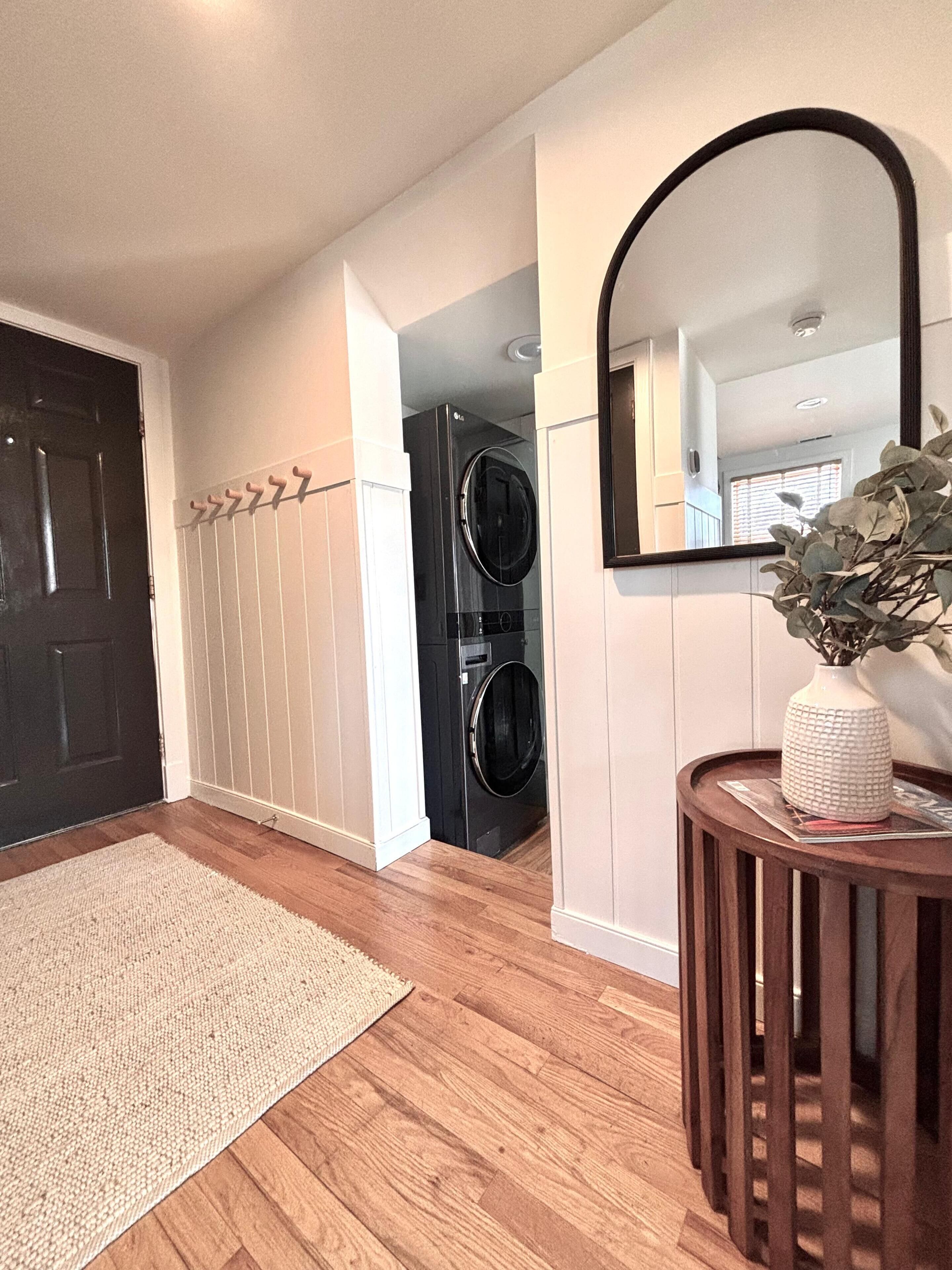 A hallway with a wooden floor, a white rug, a black door, a coat rack, a black washing machine, a mirror, and a vase with a plant.