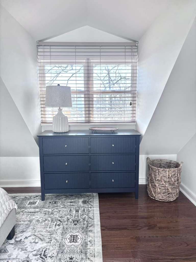 A bedroom with a large window, a dresser, a lamp, a rug, and a wicker basket.
