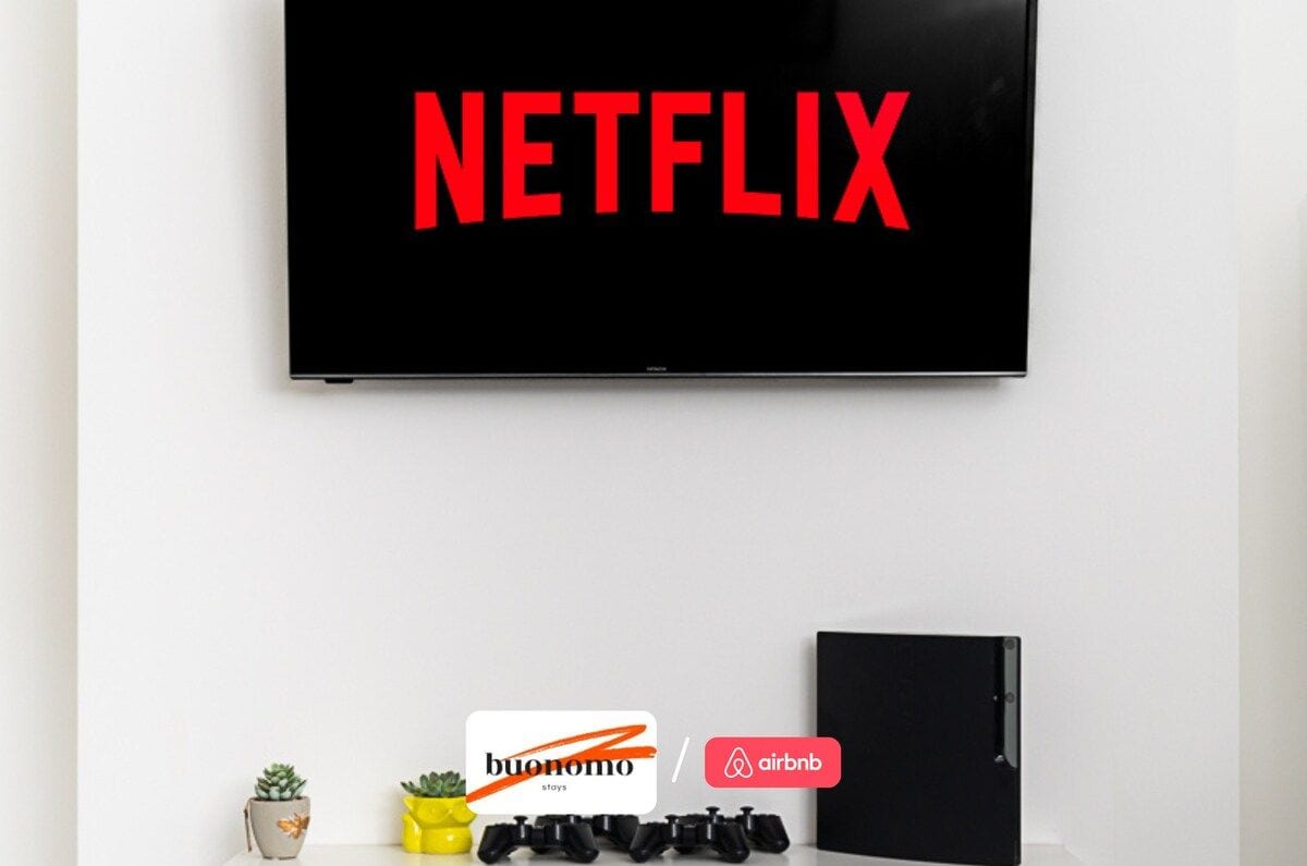 A television mounted on a white wall displaying the Netflix logo. Below the TV, there are two small potted plants, a set of black game controllers, and a black notebook or folder.