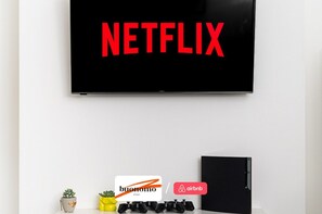 A television mounted on a white wall displaying the Netflix logo. Below the TV, there are two small potted plants, a set of black game controllers, and a black notebook or folder. - 10% OFF Cosy 3BR Flat • Easy Central London • Wifi (London)