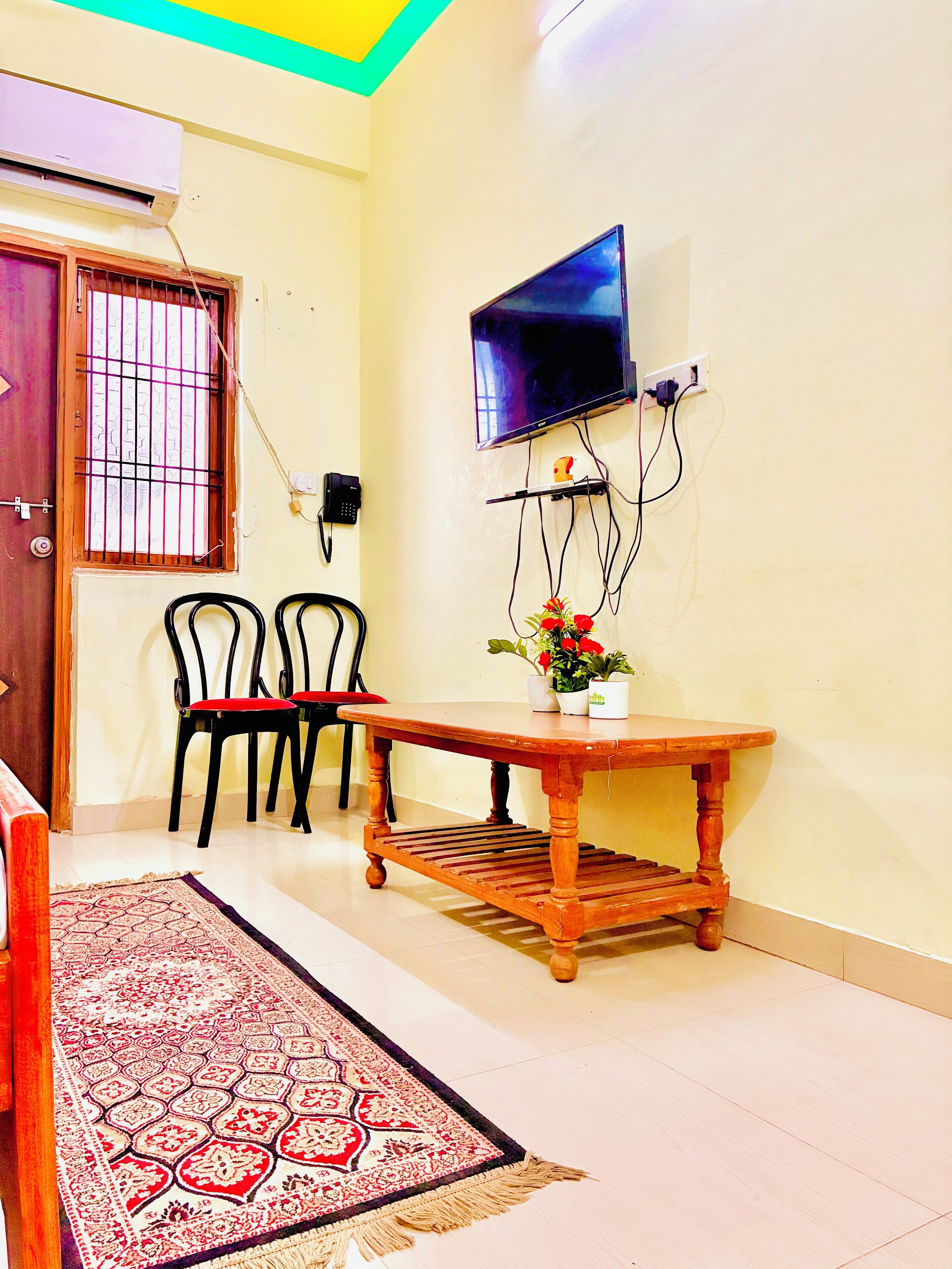 A room with a wooden table, two chairs, a TV mounted on the wall, and a patterned rug on the floor.