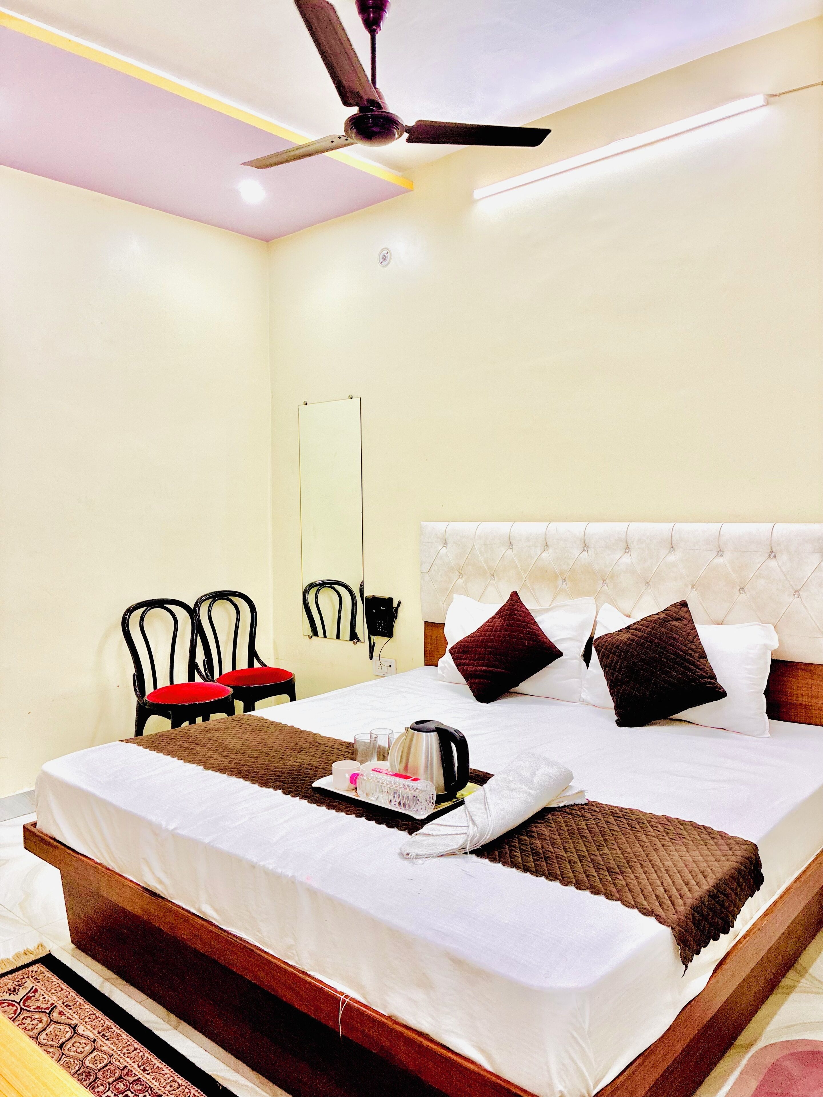 A hotel room with a bed, a ceiling fan, a bedside table with a tray of breakfast items, and two chairs.
