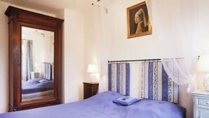 Een slaapkamer met een grote spiegel, een bed met een blauwe spreidek, een nachtkastje met een lamp en een aan de muur hangend schilderij.
