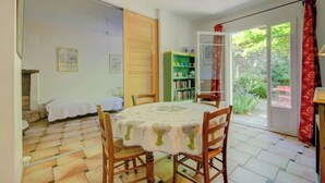Ein Essbereich mit einem runden Tisch, der mit einer Tischdecke bedeckt ist, Holzsesseln, einem Bücherregal und einer Glastür, die in einen Garten führt.