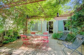 Une terrasse couverte avec une table et des chaises, entourée d’une végétation luxuriante et bordée d’un bâtiment doté d’une porte.