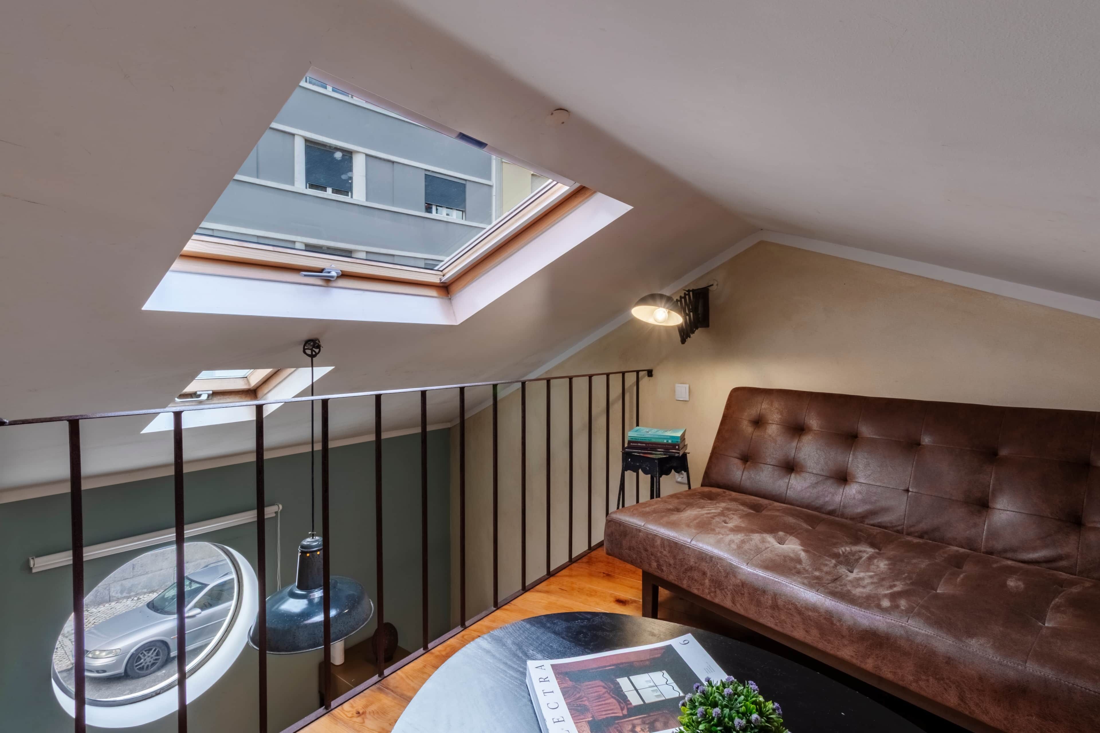 Een loft-achtige kamer met een schuine plafond, een bruine leren bank, een ronde salontafel en een dakraam.