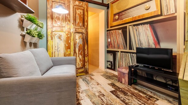 Un salon avec un canapé, un téléviseur posé sur un meuble, une bibliothèque remplie de disques et une porte dont la peinture s’écaille.