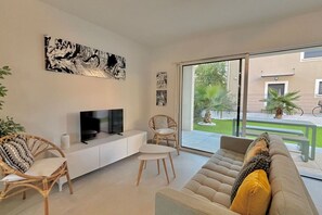 Ein modernes Wohnzimmer mit einem Flachbildfernseher auf einer weißen Konsole, Korbstühle, ein beiges Sofa mit Zierkissen und eine große Glasschiebetür, die zu einer Außenpatio mit einer Palme und einem Fahrrad führt.