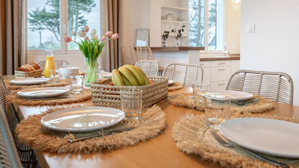 Een eettafel gedekt met borden, glazen, een fruitmand en een vaas met bloemen.
