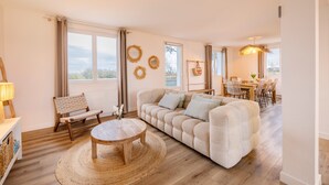 Een woonkamer met een bank, een rond houten bijzettafeltje, een rieten fauteuil en een eethoek met een tafel en stoelen.