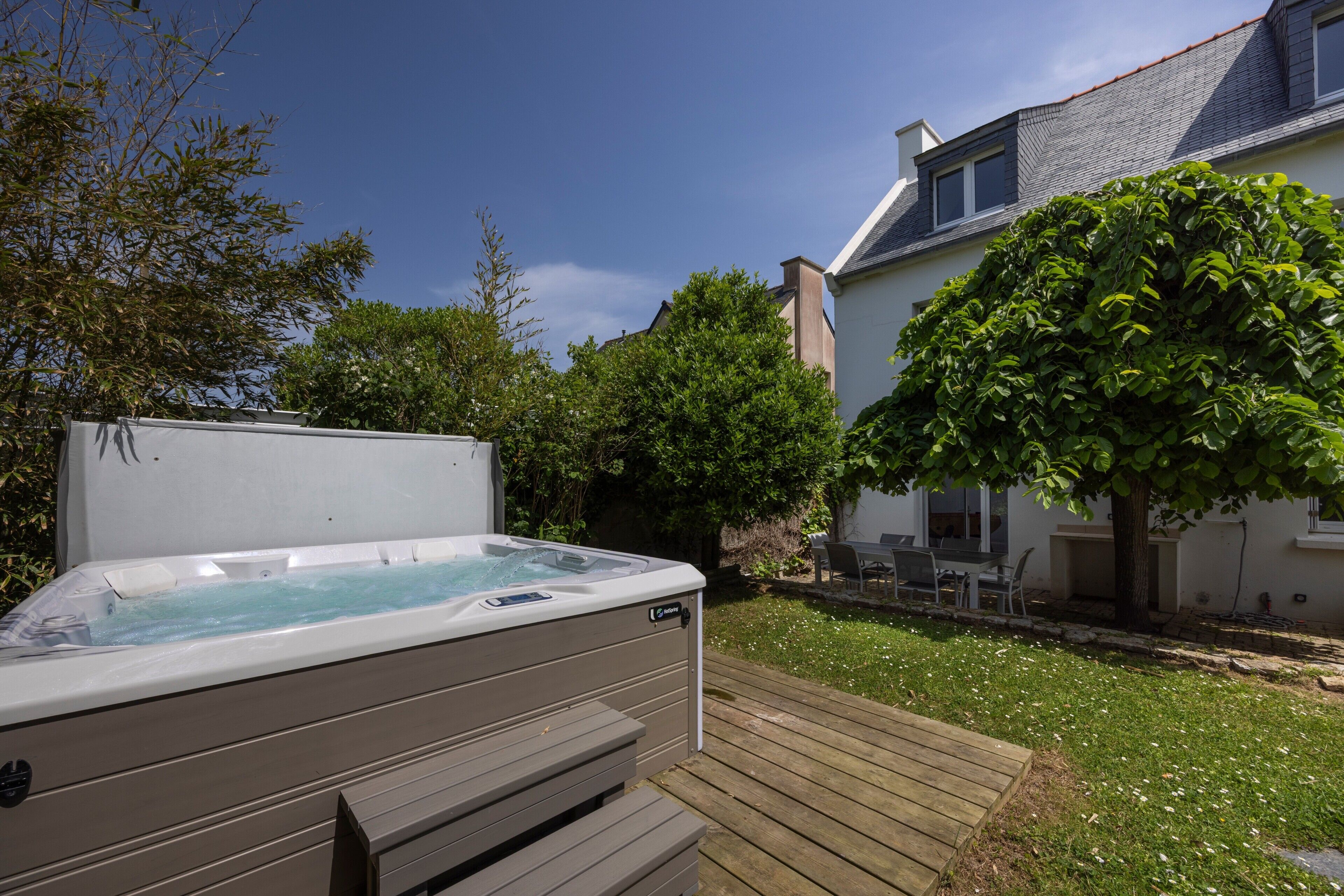 Un bain à remous dans un jardin, avec une terrasse en bois, entouré d’arbres et d’une maison.