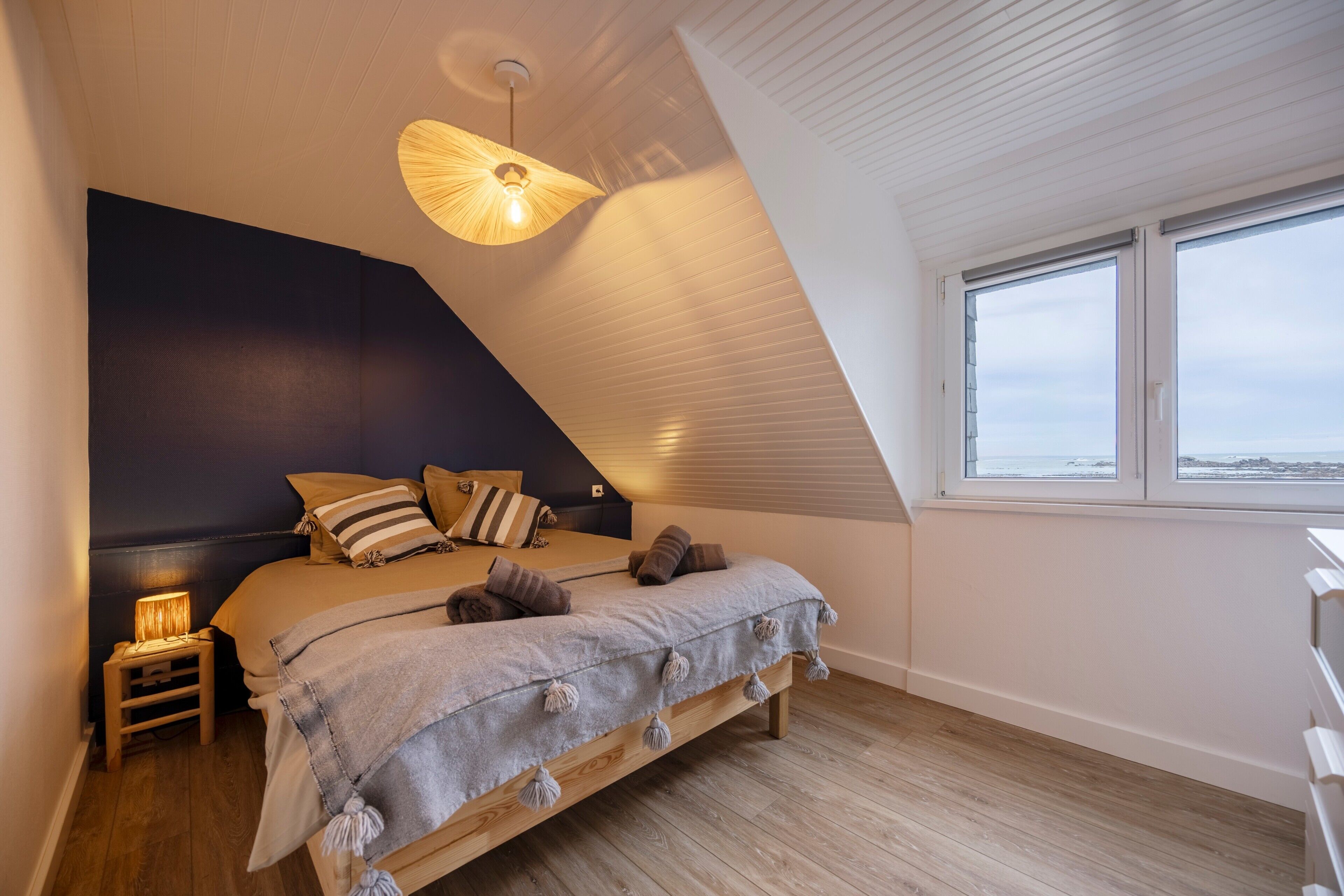 Un dormitorio con techo inclinado, una cama con manta y almohadas grises, una lámpara de noche y una ventana con vistas.