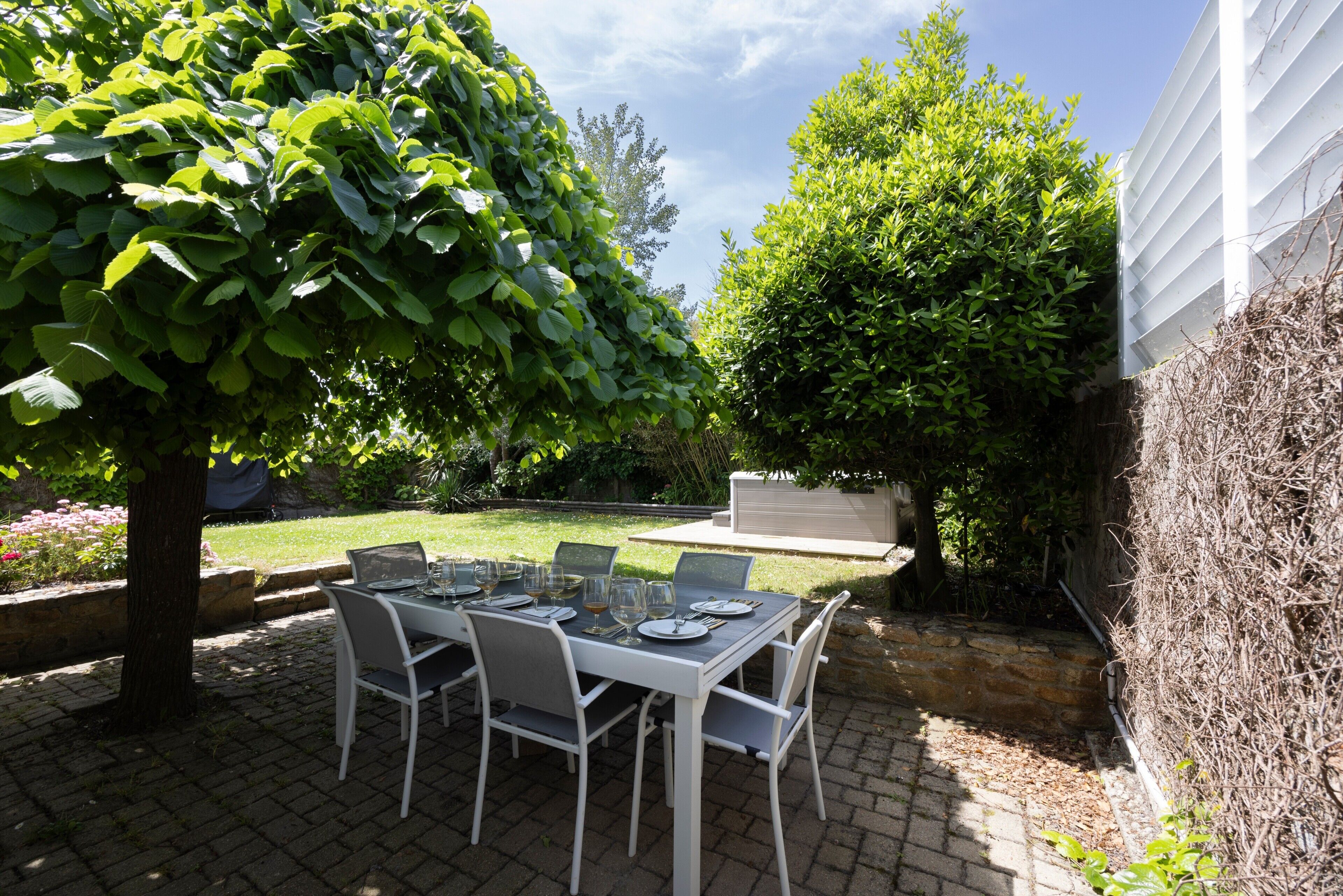 An outdoor patio with a table set for four, surrounded by chairs, a stone wall, and greenery.