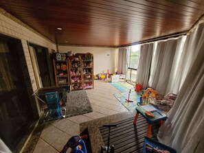 A room with a tiled floor, a wooden ceiling, a bookshelf filled with toys, a small table with a mat, and a window with curtains. - Carnival in Rio, 30 minutes from Copacabana, right in front of the beach. (Rio de Janeiro)
