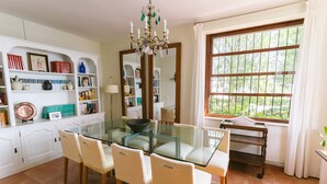 A dining area with a glass table, four chairs, a chandelier, a large window with a view of greenery, and a white cabinet with shelves. - Seafront Chalet with Pool, Outdoor Kitchen, Spacious Garden and Stunning Views (Alicante (Alacant))
