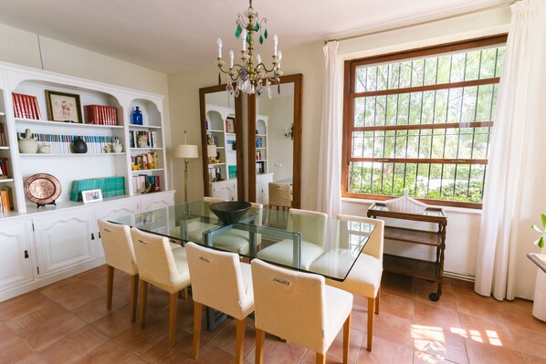 A dining area with a glass table, four chairs, a chandelier, a large window with a view of greenery, and a white cabinet with shelves.