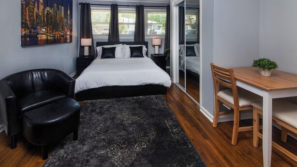 Um quarto com cama, escrivaninha, cadeira, poltrona de couro preto, tapete preto e ventilador de teto.
