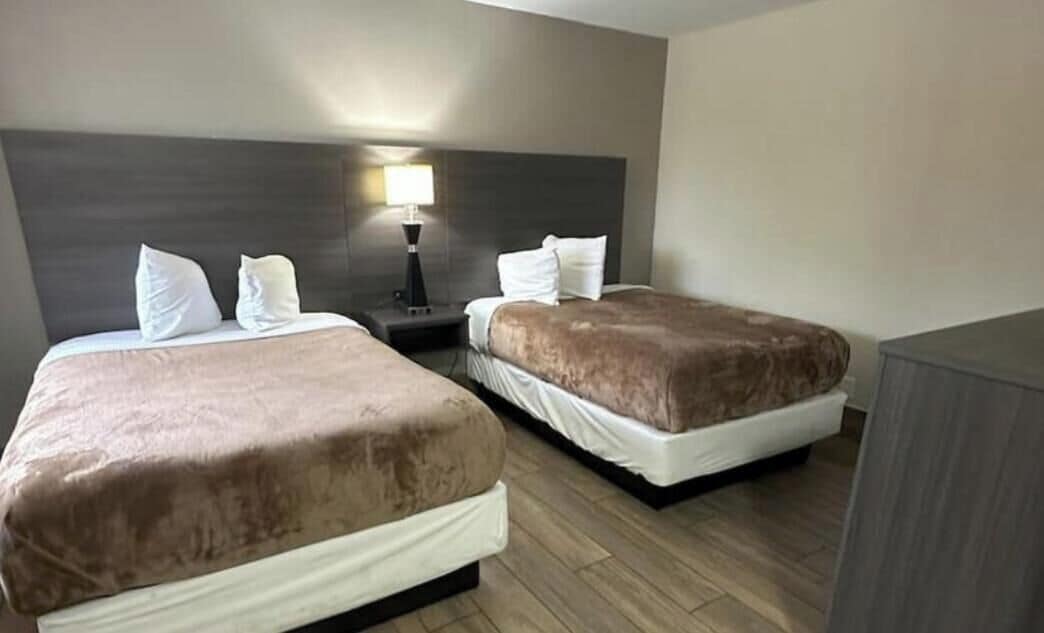 Quarto de hotel com duas camas, uma mesa de cabeceira com abajur e piso de madeira.