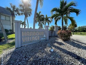 Schild für Harbour Village mit einer Grafik eines Segelboots, umgeben von Palmen und einer Schotterzufahrt.