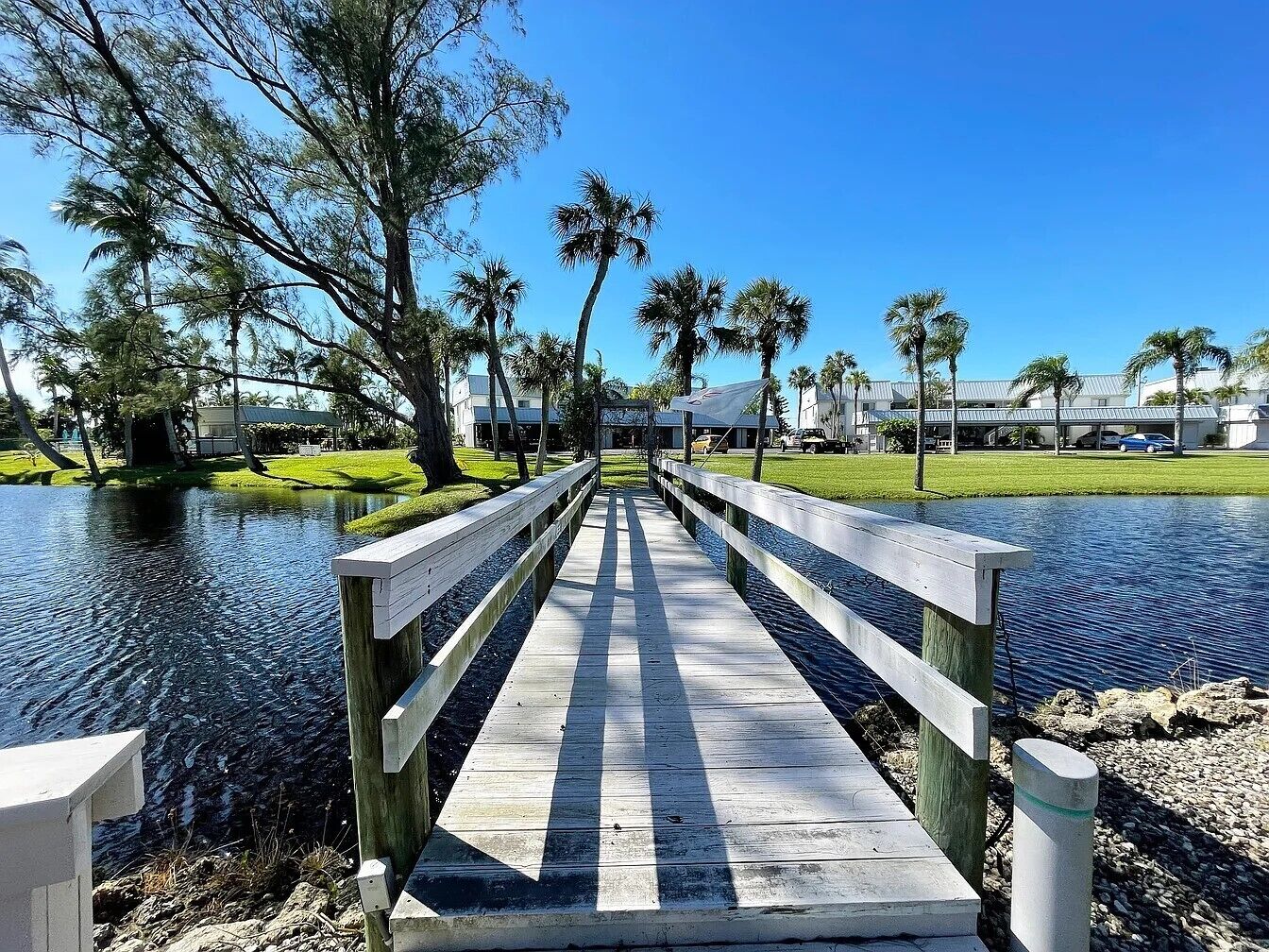 A wooden bridge over a calm water body, surrounded by trees and a grassy area.