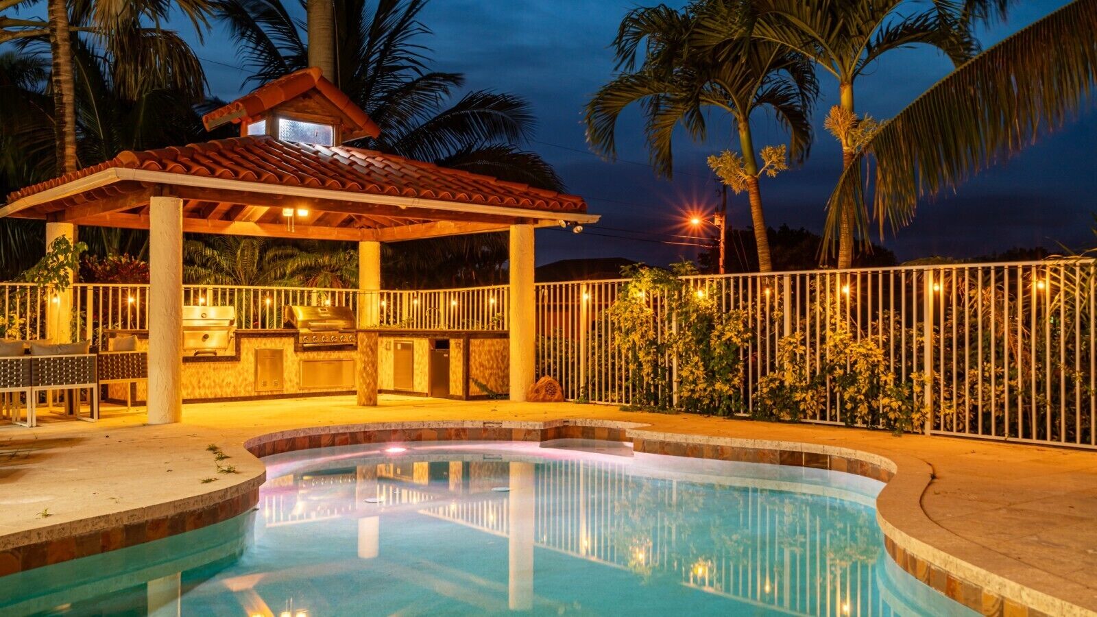 A swimming pool with a covered barbecue area, surrounded by a fence and palm trees.