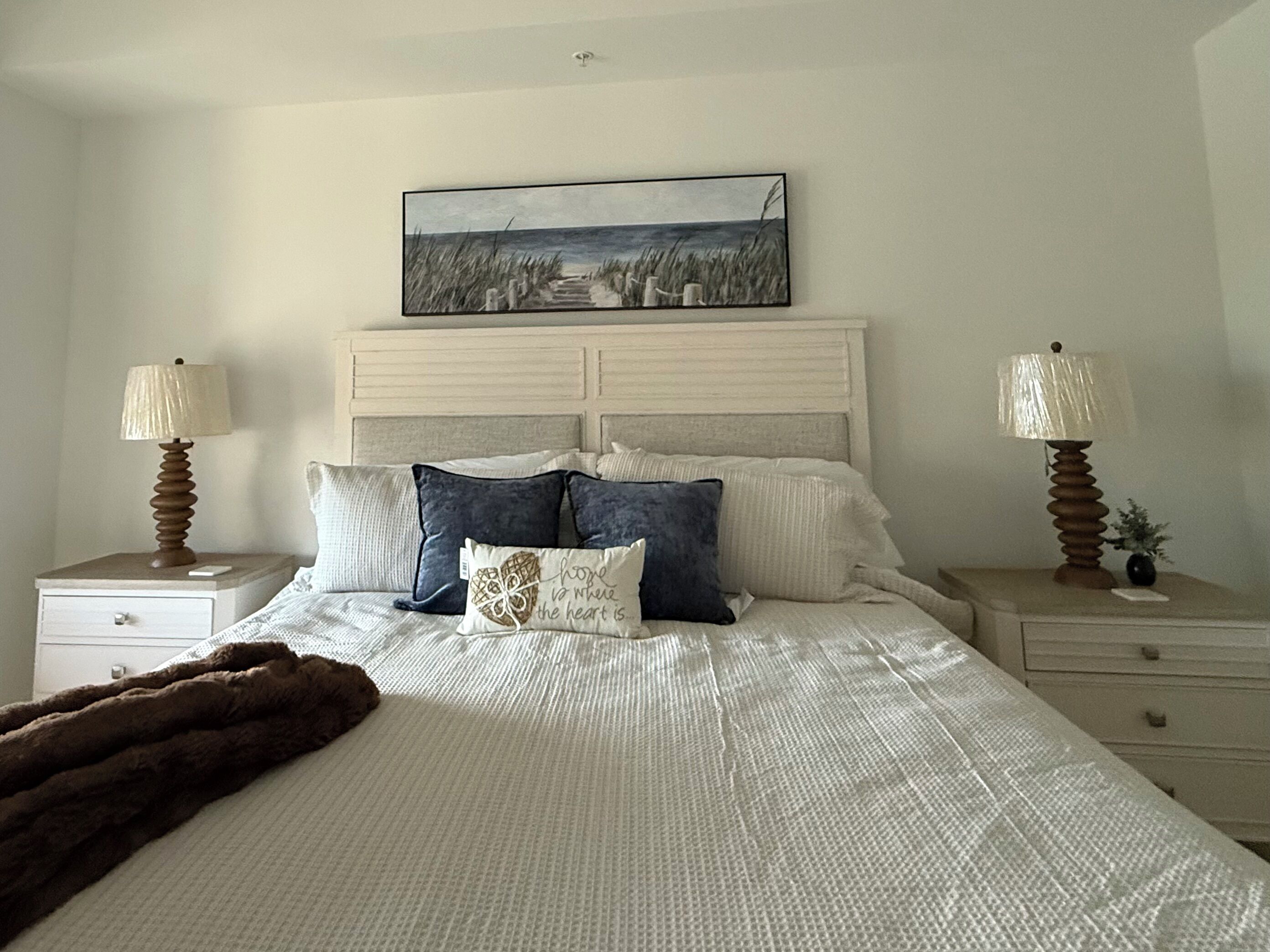Une chambre à coucher comprenant un lit, deux tables de chevet, une commode, une lampe, un tableau et un coussin décoratif.