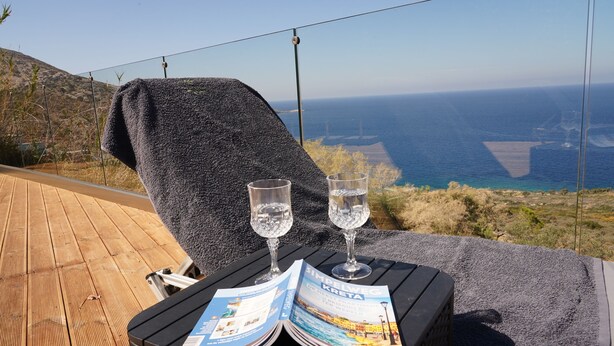 A lounge chair with a towel, two glasses of water, and a travel magazine on a wooden deck overlooking the ocean. - House of Harmony Suites (Malevizi)