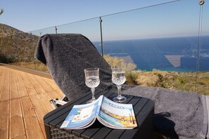 Ein Liegestuhl mit Handtuch, zwei Gläsern Wasser und einer Reisemagazin auf einem Holzdeck mit Blick aufs Meer.
