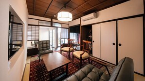 Il s’agit d’une pièce de style japonais traditionnel, avec un sol recouvert de tatamis, un plafond bas en bois et des portes coulissantes. On y trouve un canapé, une table et deux chaises.