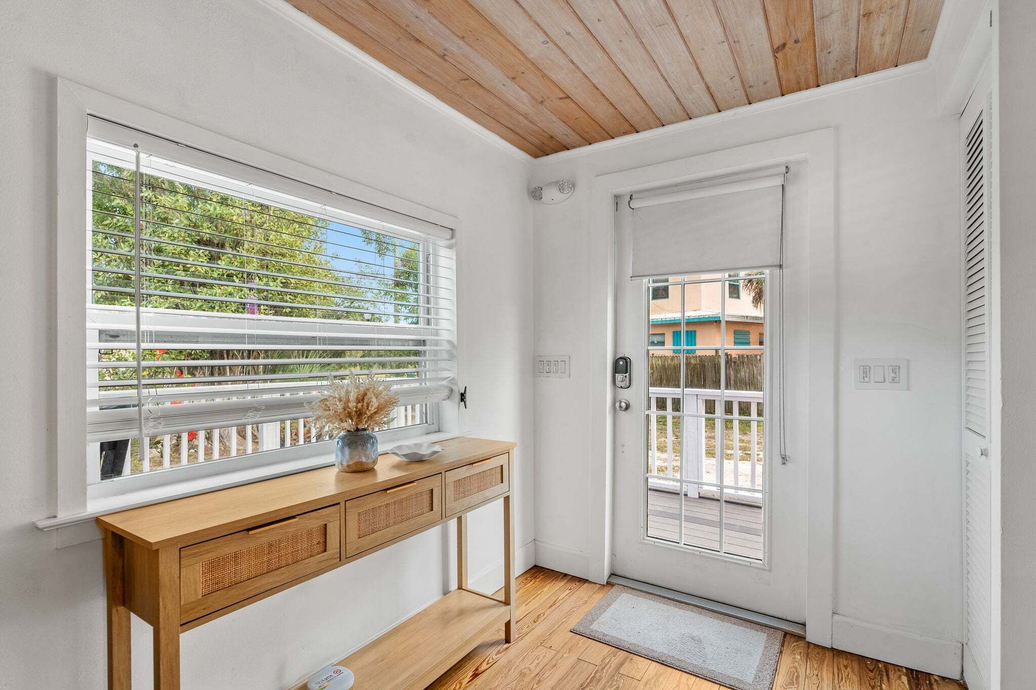 Una stanza con soffitto in legno, una porta bianca con un vetro, un consolle in legno con cassetti e una finestra con persiane.