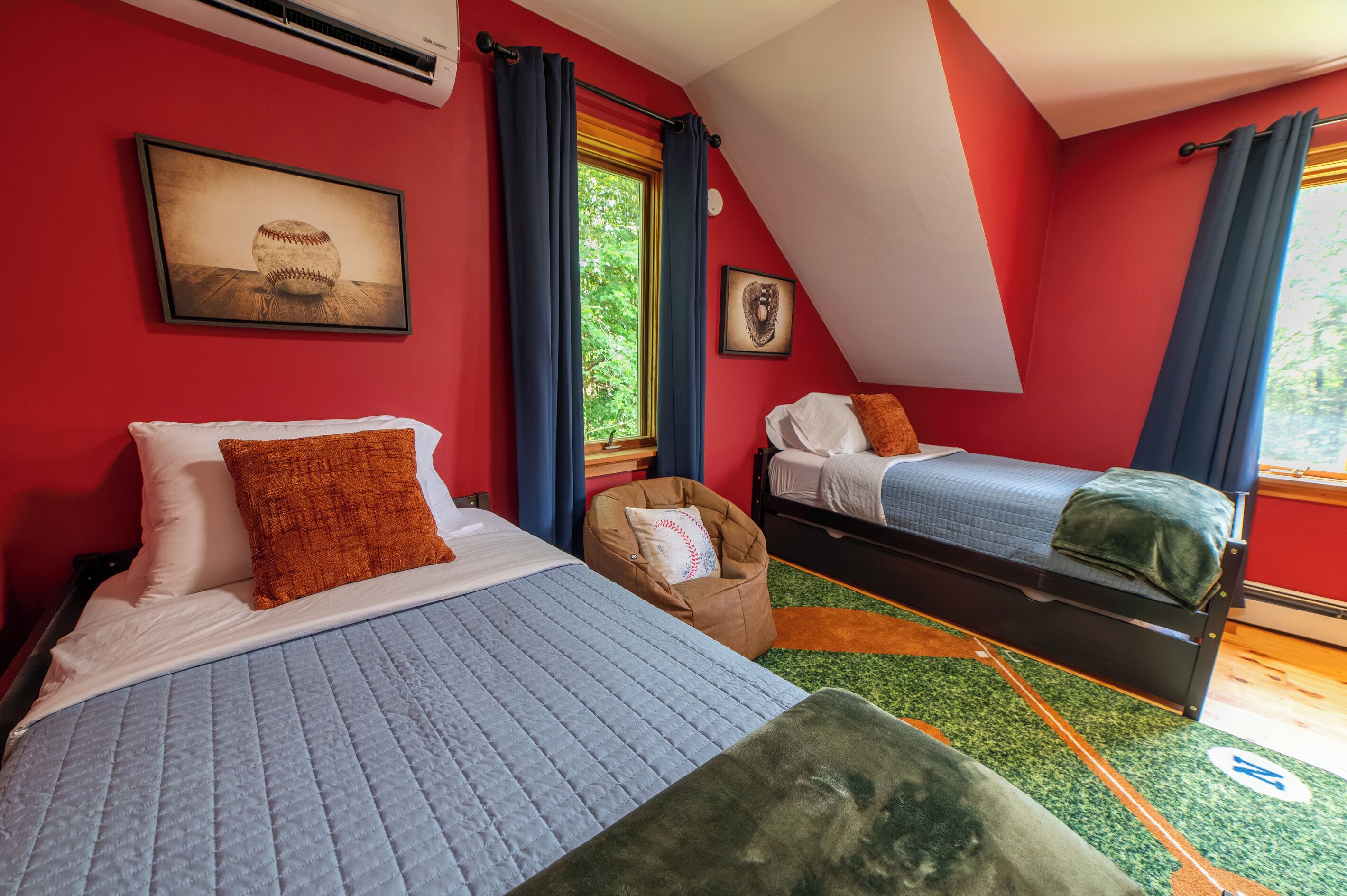 A room with two beds, a window with curtains, a framed picture on the wall, and a small chair with a baseball pillow.