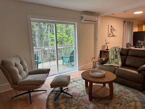 Un salon comprenant un canapé, un fauteuil et un pouf, une petite table ronde et une porte-fenêtre coulissante donnant sur une terrasse extérieure.