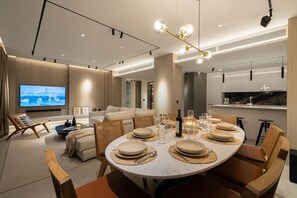 Ein moderner Essbereich mit einem runden Tisch, gedeckt für sechs Personen, einem an der Wand befestigten Fernseher und einem sichtbaren Küchenbereich im Hintergrund.