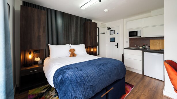 Un dormitorio con cama, mesita de noche, lámpara y un osito de peluche. Hay una kitchenette con microondas y armarios.