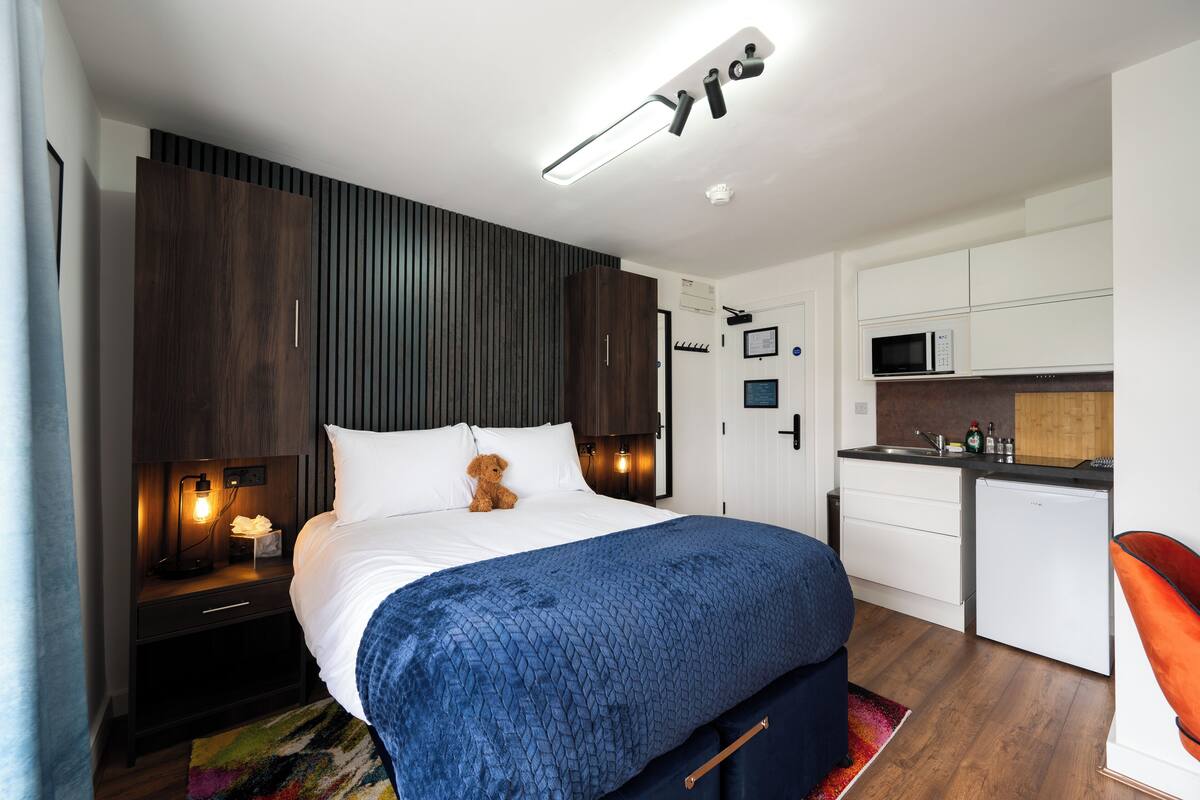 A bedroom with a bed, bedside table, lamp, and a teddy bear. There is a kitchenette with a microwave and cabinets.