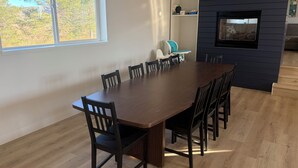 A dining area with a large wooden table and chairs, a window with a view of trees, and a dark-colored wall with a built-in TV.
