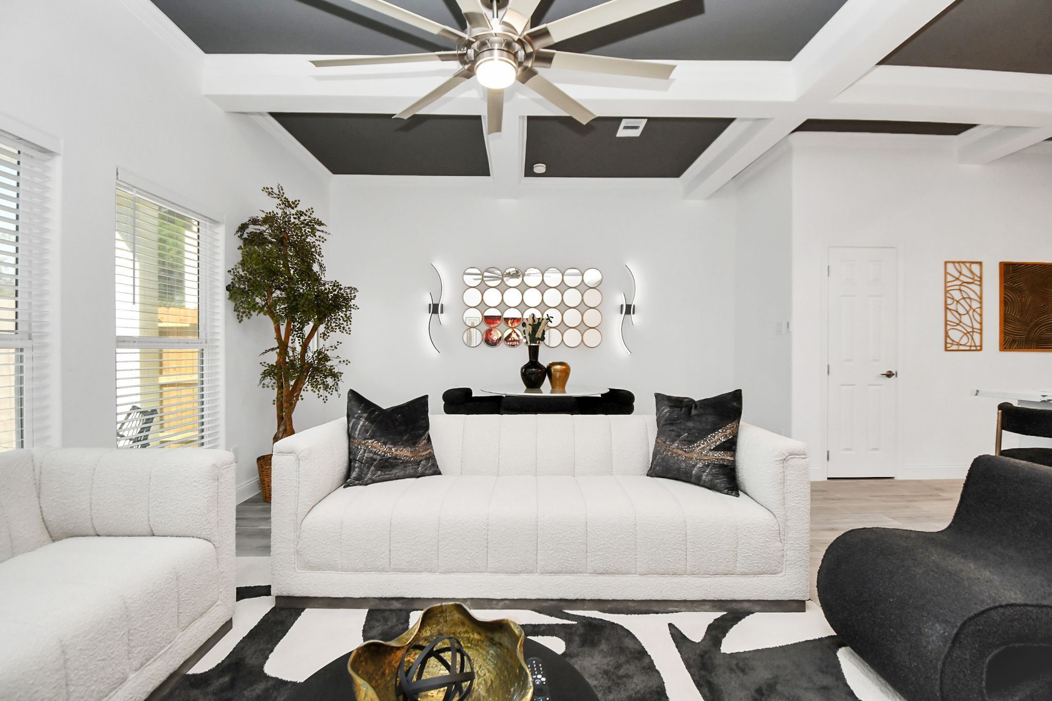 A modern living room with a white sofa, black and white rug, ceiling fan, and wall-mounted lighting.