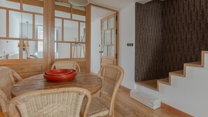 Un comedor con mesa de madera y sillas de mimbre, un cuenco rojo sobre la mesa y una escalera con una pared oscura detrás.