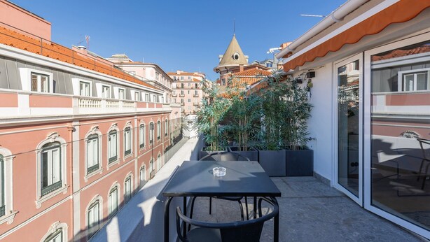 Un balcon avec une table et des chaises, donnant sur un paysage urbain composé de bâtiments traditionnels et du clocher d’une église.