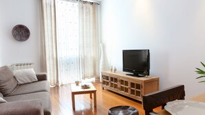 Een woonkamer met een bank, een salontafel, een televisie op een houten kast en een raam met gordijnen.