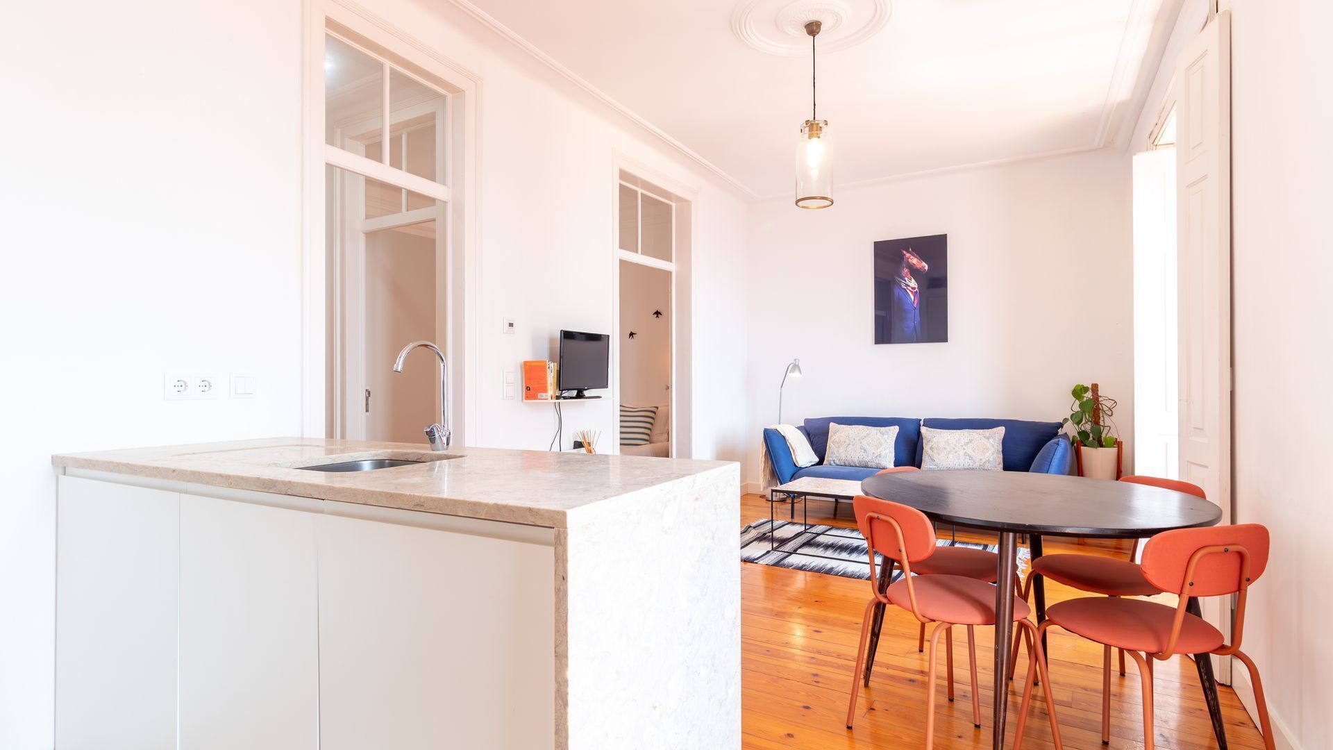 A modern kitchen with a large island, a dining area with a round table and orange chairs, and a living room with a sofa and a television.